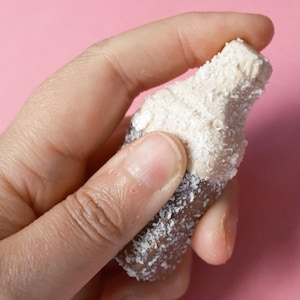 Könnte beinhalten: Ein kleines, flaschenförmiges Bonbon mit brauner Basis und hellbeigem Oberteil, beide mit einer feinen Schicht aus weißen Kokosraspeln bedeckt. Das Konfekt hat eine ausgeprägte Hals- und Verschlussform, die an eine Miniatur-Getränkeflasche erinnert.