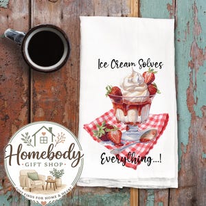 May include: White kitchen towel with a watercolor illustration of an ice cream sundae with strawberries and whipped cream. Text reads "Ice Cream Solves Everything...!". A black coffee mug and a Homebody Gift Shop logo are also visible.