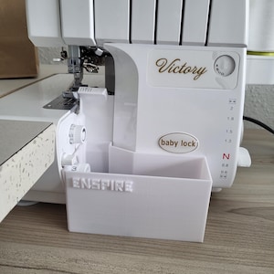 May include: A white Baby Lock Victory serger sewing machine. The machine has the word "Victory" in gold script and the word "Enspire" on the removable waste bin. The machine is on a wooden surface.