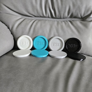 May include: A selection of 3D-printed circular lids in white, light blue, grey, and black. Each lid features the embossed text "T3D". The lids are displayed on a grey leather surface.