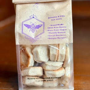 May include: A package of honey shortbread cookies in a clear plastic bag, inside a brown paper bag. The paper bag has a purple bee logo and text that reads "Johnny & Elle Est 2015" and "Hand Made With Raw Honey". The cookies weigh 7 oz.