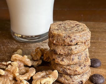 May include: A stack of six square-shaped cookies with visible chocolate chips and nuts. Beside the cookies are walnut halves and dark chocolate discs. A glass of milk is in the background, all on a wooden surface.
