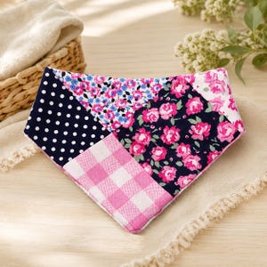Peut inclure: Un bandana triangulaire pour animal de compagnie présentant un motif patchwork composé de quatre tissus distincts : bleu marine à pois blancs, bleu clair à petites fleurs roses, bleu marine foncé à roses fuchsia et un motif vichy rose et blanc.