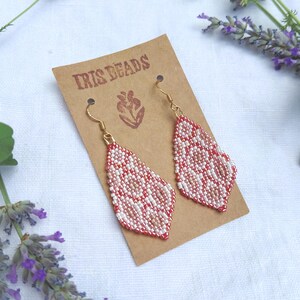May include: A pair of diamond-shaped beaded earrings featuring a geometric pattern in shades of red, pink, and white. The earrings are attached to a brown cardboard display card labeled with the text IRIS BEADS and a small flower icon.
