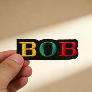 Könnte beinhalten: Gestickter Aufnäher mit dem Namen "BOB" in gelben, grünen und roten Buchstaben auf schwarzem Grund. Der Aufnäher ist rechteckig mit gewelltem Rand. Die Buchstaben sind mit dickem Faden gestickt.