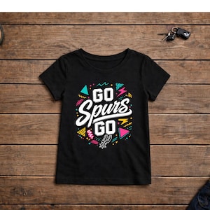 May include: Black t-shirt with the text "GO Spurs GO" in white, with colorful geometric shapes. The shirt has a crew neck and short sleeves. The design includes teal, pink, yellow, and black accents. The shirt is displayed on a wooden surface.