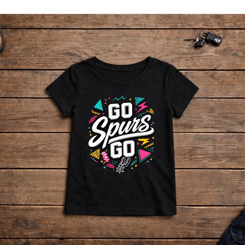 May include: Black t-shirt with the text "GO Spurs GO" in white, with colorful geometric shapes. The shirt has a crew neck and short sleeves. The design includes teal, pink, yellow, and black accents. The shirt is displayed on a wooden surface.
