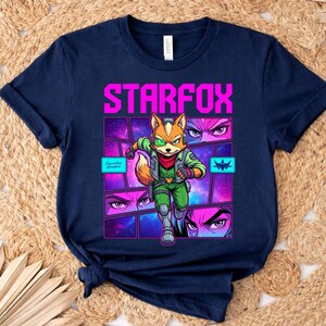 May include: A navy blue t-shirt featuring a graphic print of the character Fox McCloud from Starfox. The design includes the text STARFOX in bright pink, a central illustration of the character, and smaller panels showing close-ups of eyes.
