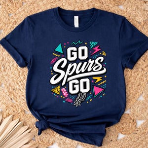 May include: Navy blue t-shirt with the text "GO Spurs GO" in white, outlined in black. The design includes colorful geometric shapes and the Spurs logo. The shirt is folded on a woven surface.