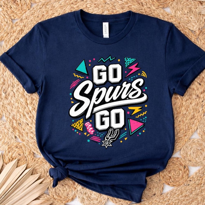 May include: Navy blue t-shirt with the text "GO Spurs GO" in white, outlined in black. The design includes colorful geometric shapes and the Spurs logo. The shirt is folded on a woven surface.