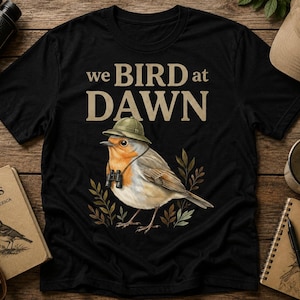 Könnte beinhalten: Ein schwarzes Kurzarm-T-Shirt mit einer Grafik eines Rotkehlchens, das einen Tropenhelm und ein Fernglas trägt. Der Text über dem Vogel lautet "we BIRD at DAWN" in beiger Schrift. Das Design enthält zudem kleine grüne und braune Blätter.