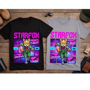May include: A black t-shirt and a gray t-shirt, each featuring a graphic print of the character Fox McCloud from Starfox. The design includes the text STARFOX in bright pink, a space-themed background with comic-style panels, and a green visor.