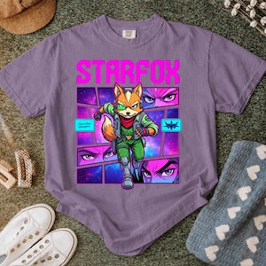 May include: A purple short-sleeve t-shirt featuring a graphic print of the character Fox McCloud from Star Fox. The design includes the text STARFOX in bold pink letters, a central character illustration, and smaller panels showing eyes.