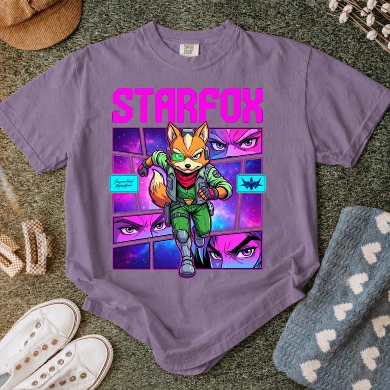 May include: A purple short-sleeve t-shirt featuring a graphic print of the character Fox McCloud from Star Fox. The design includes the text STARFOX in bold pink letters, a central character illustration, and smaller panels showing eyes.