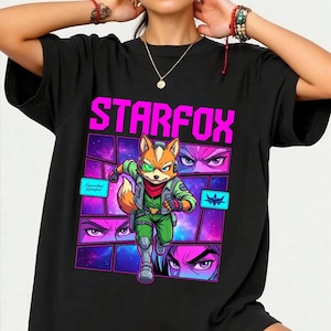 May include: A black short-sleeved t-shirt featuring a graphic print with the text STARFOX in bright pink. The design includes a central illustration of a fox character in a green flight suit, surrounded by comic-style panels with space imagery.