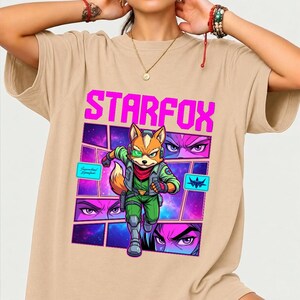 May include: A beige short-sleeved t-shirt featuring a graphic print of the character Fox McCloud from Star Fox. The design includes the text STARFOX in bright pink, a central character illustration, and smaller panels showing eyes and a spaceship.