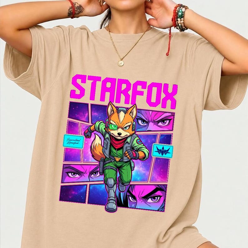 May include: A beige short-sleeved t-shirt featuring a graphic print of the character Fox McCloud from Star Fox. The design includes the text STARFOX in bright pink, a central character illustration, and smaller panels showing eyes and a spaceship.