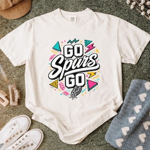 May include: White t-shirt with the text "GO Spurs GO" in a colorful, retro design. The design includes geometric shapes, lightning bolts, and the Spurs logo. A casual tee for fans.