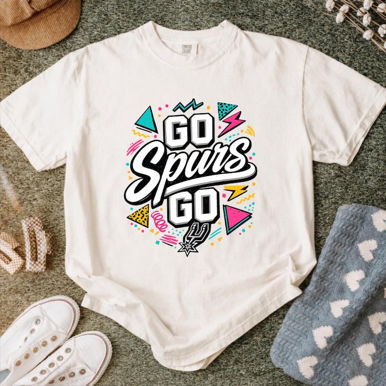 May include: White t-shirt with the text "GO Spurs GO" in a colorful, retro design. The design includes geometric shapes, lightning bolts, and the Spurs logo. A casual tee for fans.