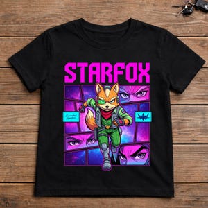 May include: A black short-sleeved t-shirt featuring a graphic print of a fox character in a green flight suit with the text STARFOX in bright pink. The design includes a cosmic purple background, segmented comic-style panels, and close-up eye details.