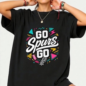 May include: Black t-shirt with the text "GO Spurs GO" in white, outlined in black. The design includes colorful geometric shapes and the Spurs logo. The shirt has short sleeves and a crew neck.