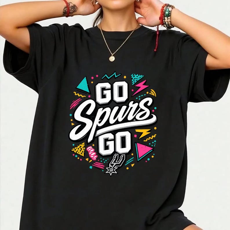 May include: Black t-shirt with the text "GO Spurs GO" in white, outlined in black. The design includes colorful geometric shapes and the Spurs logo. The shirt has short sleeves and a crew neck.
