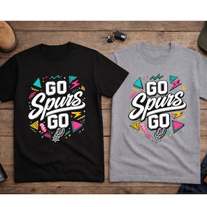 May include: Two t-shirts, one black and one gray, with the text "GO Spurs GO" in a colorful, retro design. The design includes geometric shapes and the Spurs logo. The shirts are displayed on a wooden surface.