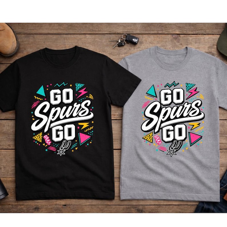 May include: Two t-shirts, one black and one gray, with the text "GO Spurs GO" in a colorful, retro design. The design includes geometric shapes and the Spurs logo. The shirts are displayed on a wooden surface.