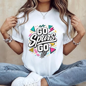 May include: White t-shirt with the text "GO Spurs GO" in a bold, stylized font. The design includes colorful geometric shapes and the Spurs logo. The shirt is a classic crew neck style.
