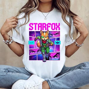 May include: A white t-shirt featuring a graphic print of the character Fox McCloud from Starfox. The design includes the text STARFOX in bright pink, a central illustration of the character in a green flight suit, and surrounding comic panels.