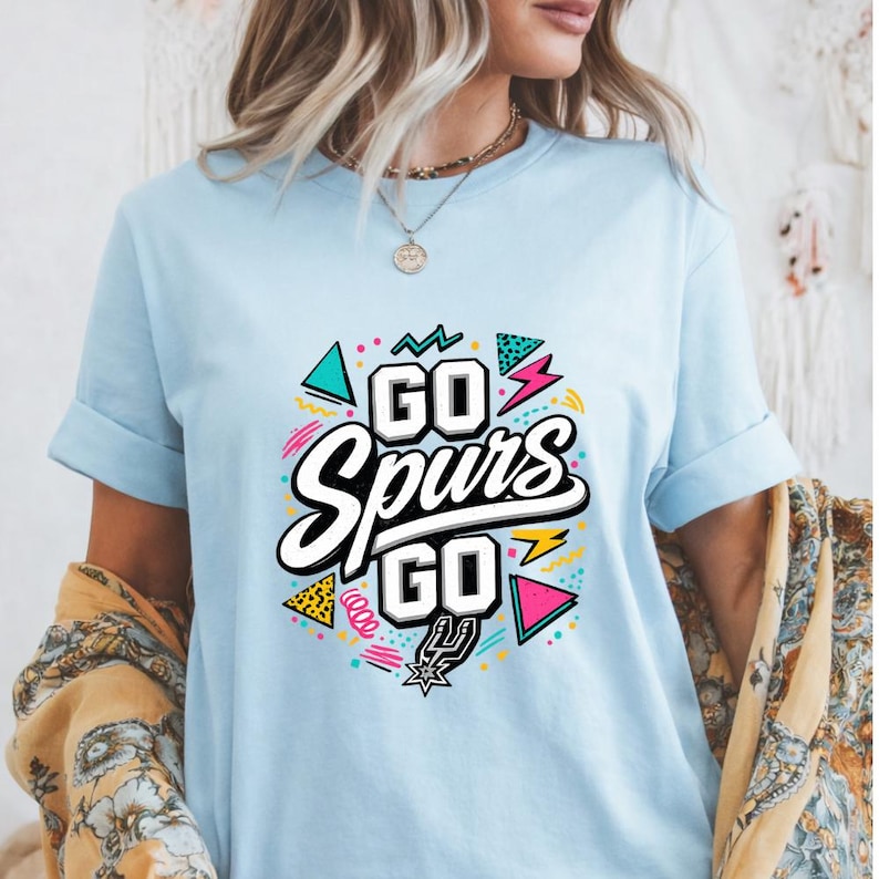 May include: Light blue t-shirt with the text "GO Spurs GO" in a colorful design. The design includes geometric shapes, lightning bolts, and the Spurs logo. The shirt has short sleeves and a crew neck.
