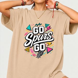 May include: A tan t-shirt with the words "GO Spurs GO" in a colorful graphic design. The design includes geometric shapes, lightning bolts, and the Spurs logo. The shirt is a casual, comfortable style.