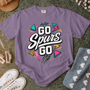 May include: A purple t-shirt with the text "GO Spurs GO" in a colorful design. The design includes geometric shapes in teal, pink, and yellow, with a black and white outline. The shirt is made of a soft material.