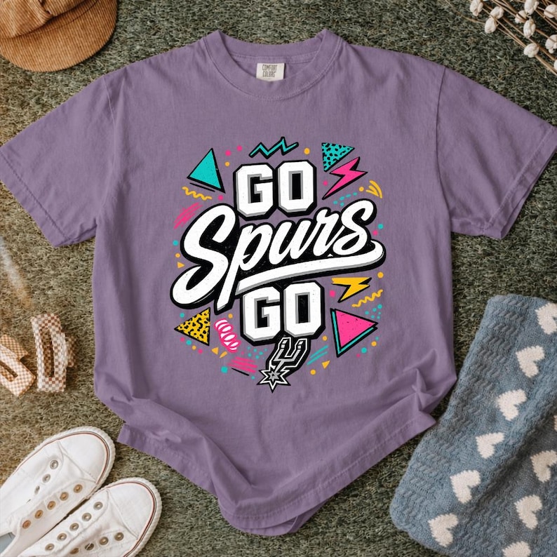 May include: A purple t-shirt with the text "GO Spurs GO" in a colorful design. The design includes geometric shapes in teal, pink, and yellow, with a black and white outline. The shirt is made of a soft material.