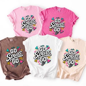 May include: Five t-shirts in various colors: pink, hot pink, brown, white, and beige. Each shirt features the text "GO Spurs GO" in a bold, stylized font with colorful geometric accents and a Spurs logo.