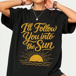 以下が含まれることがあります： 黒の半袖Tシャツで、筆記体の黄色い文字で「I'll Follow You into the Sun」とプリントされています。テキストの下には、太陽の光と水を表現した波線が描かれた、昇る太陽のグラフィックが黄色でデザインされています。