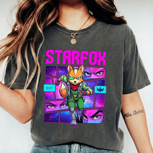 May include: A charcoal gray t-shirt featuring a graphic print of the character Fox McCloud from Star Fox. The design includes the text STARFOX in bright pink, a space-themed background with purple and blue hues, and comic-style panels.