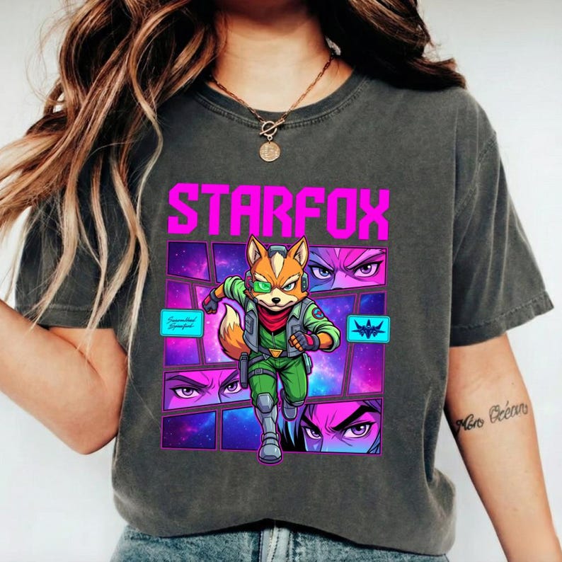 May include: A charcoal gray t-shirt featuring a graphic print of the character Fox McCloud from Star Fox. The design includes the text STARFOX in bright pink, a space-themed background with purple and blue hues, and comic-style panels.