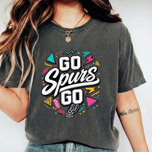 May include: Dark gray t-shirt with the text "GO Spurs GO" in a white, stylized font, surrounded by colorful geometric shapes and lightning bolts. The San Antonio Spurs logo is at the bottom. A casual, comfortable tee.