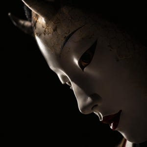 以下が含まれることがあります： 鋭い角、暗いアーモンド形の目、光沢のある赤い唇が特徴の白い陶器製の般若の面。額の部分には質感のある金色のひび割れ模様が施され、黒い髪は後ろにまとめられており、背景は無地の黒色となっている。