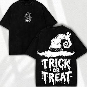 May include: A black short-sleeve t-shirt featuring a white graphic of a winking ghost and the text BOO on the front. The back displays a large white witch hat with a star-patterned band, a small cat, and the text TRICK OR TREAT in a dripping font.