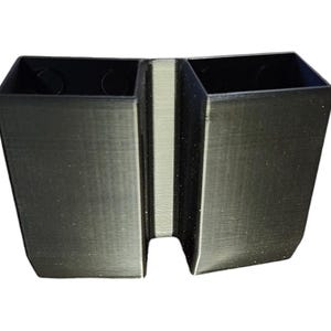 May include: A black, dual-compartment organizer with a textured surface. The organizer has two rectangular sections joined by a central divider. The top edges of the compartments are open, and the base has a slight inward angle.