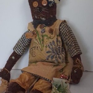 Könnte beinhalten: Handgemachte Folk-Art-Puppe mit braunem Kopf, Knopfaugen und einer blau-weiß gemusterten Schleife. Die Puppe trägt ein gelbes Kleid mit Blumenmuster über einem karierten Hemd und braunen Hosen. Ein kleiner Strumpf mit Blumenmuster liegt zu ihren Füßen.