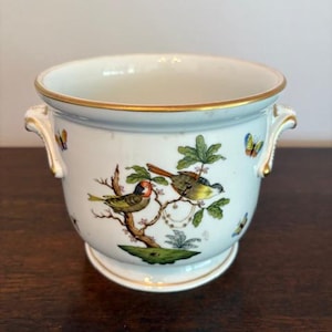 Może przedstawiać: Biała ceramiczna donica ze złotą obwódką i uchwytami. Donica posiada malowany wzór ptaków siedzących na gałęzi z zielonymi liśćmi i kolorowymi motylami. Donica jest przedmiotem dekoracyjnym.