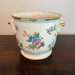 May include: A white ceramic planter with a gold rim and handles. The planter is decorated with colourful floral designs in pink, blue, and green. The planter has a round shape and sits on a small base.