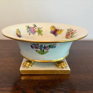 May include: A decorative porcelain bowl with a light blue exterior and a white interior. The bowl features a gold rim and is adorned with colorful butterfly and floral designs. It rests on a square base with gold-colored claw feet.