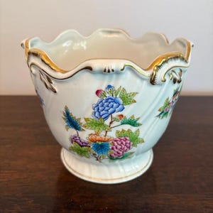 Puede incluir: Un cuenco de porcelana blanca con borde festoneado y base de pedestal. El cuenco está decorado con coloridos estampados florales, incluyendo flores azules, rosas y naranjas. Acentos dorados adornan el borde.