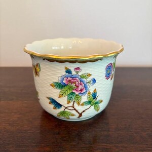 Peut inclure: Petit bol en céramique blanc cassé avec un bord festonné et une bordure dorée. Le bol est décoré de motifs floraux et de papillons colorés. L'extérieur a un aspect texturé et tissé.