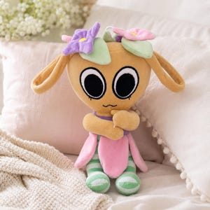 May include: A plush toy in the shape of a cartoon character with large black eyes, long ears, and a flower crown. The toy has a tan body, pink and green accents, and striped green and white legs. The toy is sitting on a bed.