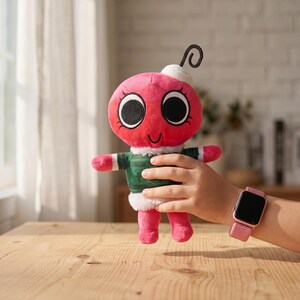 May include: A pink plush toy shaped like an ornament, featuring large black eyes, a white top, and a curly black antenna. It wears a green and white striped shirt and has pink limbs. A pink smartwatch is visible on a wrist.